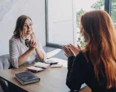 Two women talking sitting at a desk by a window