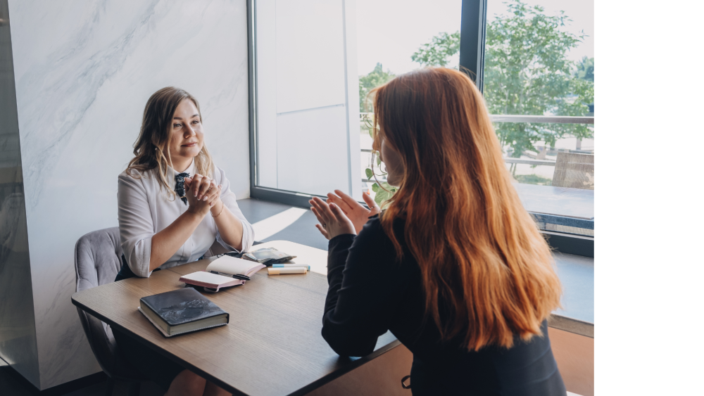 Two women talking sitting at a desk by a window